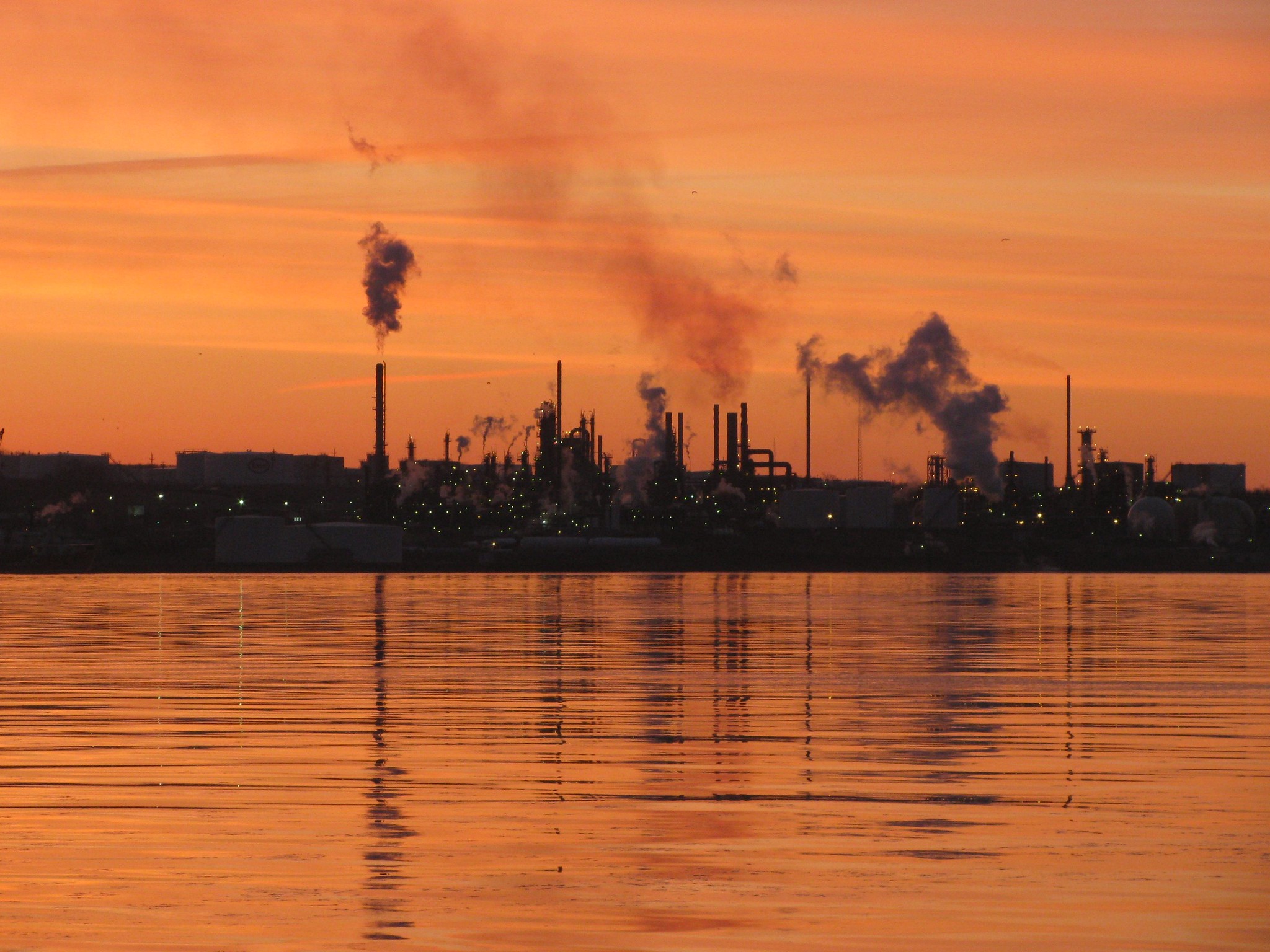 Foto van een olieraffinaderij bij zonsopgang, met rook die uit de schoorstenen komt.