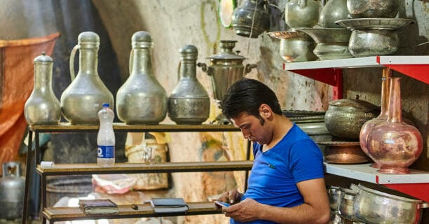 Yazd, Iran - April 22, 2017: A young man uses a smartphone while sitting on a chair in a tin shop.